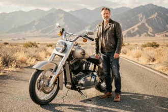 Homme en veste en cuir debout à côté d'une Harley Davidson Softail sur une route ouverte, illustrant l'assurance moto Harley Davidson